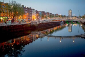 A bridge over a calm river with buildings on either side, a clear sky, and a reflection of lights on the water.