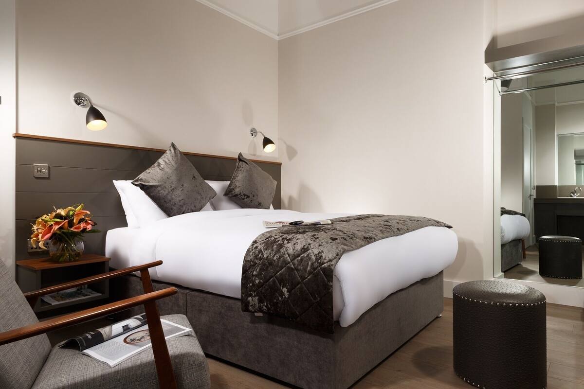 A modern hotel room with a large bed, bedside lamps, a small table with a vase of flowers, and a mirror in the corner.