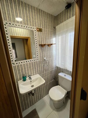 A bathroom with striped wallpaper, a sink, a toilet, a mirror, and a window with a curtain. - Hostal avenida 57 (Coslada)