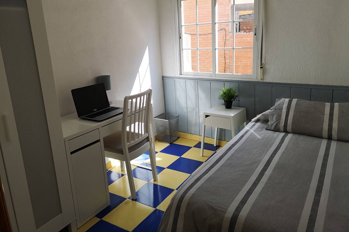 A small, neatly arranged room with a bed, a desk with a laptop, a chair, a small table with a plant, and a window with a view of a brick building.