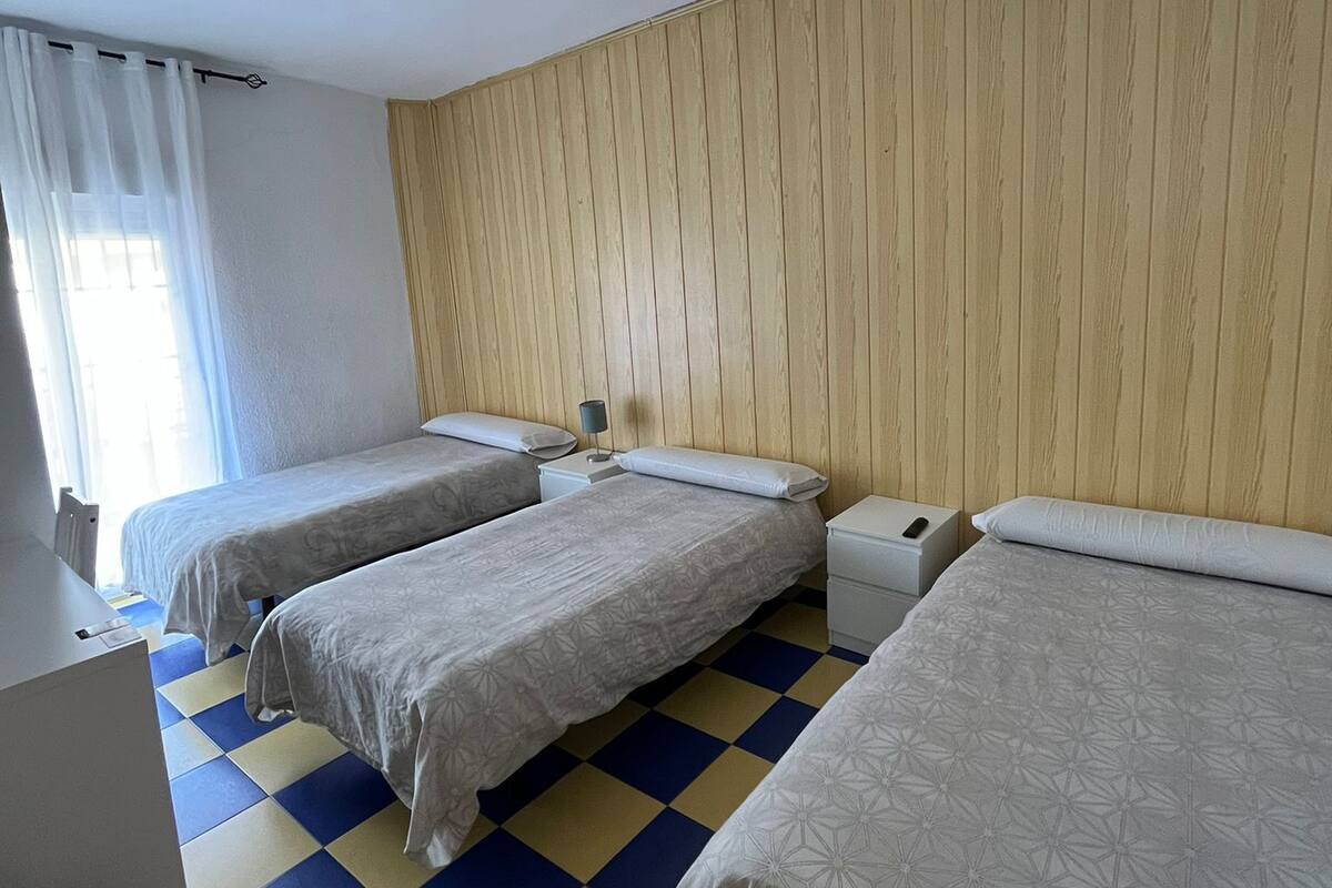A hotel room with three single beds, a wooden panel wall, a checkered floor, and a window with sheer curtains.