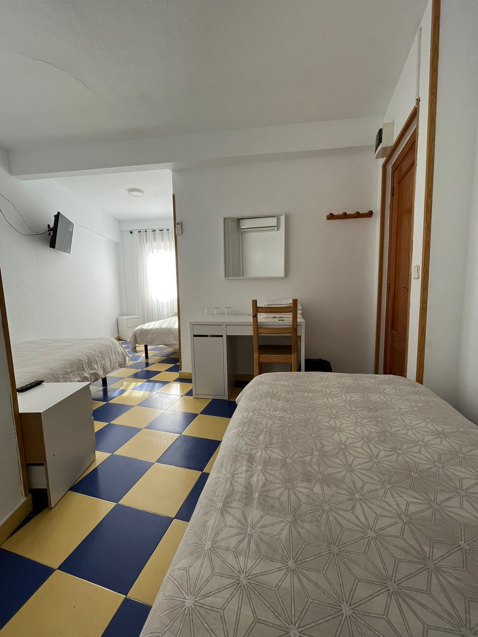A hotel room with a checkered floor, a bed, a desk, a chair, a mirror, and a television.
