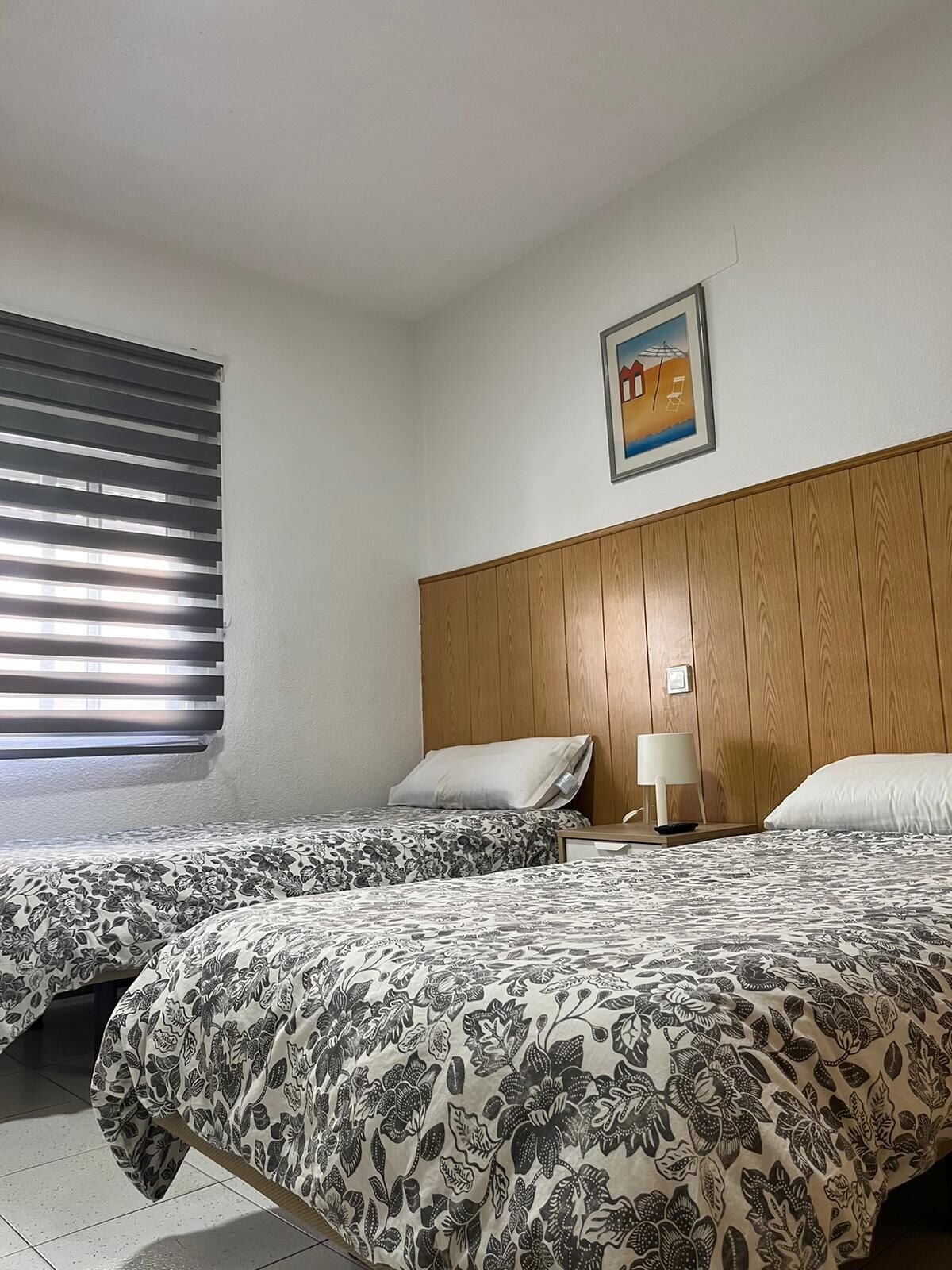 A hotel room with two beds, a wooden headboard, a window with blinds, a framed picture on the wall, and a bedside table with a lamp.