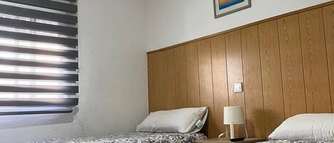 A hotel room with two beds, a wooden headboard, a window with blinds, a framed picture on the wall, and a bedside table with a lamp.