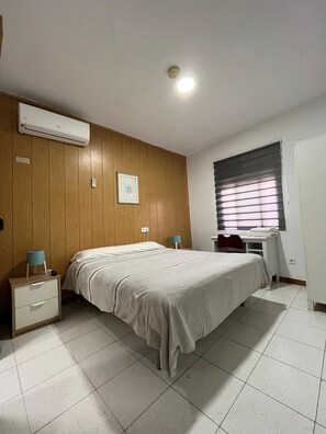 Un dormitorio con cama, mesitas de noche, escritorio, ventana con persianas, unidad de aire acondicionado y un cuadro en la pared.