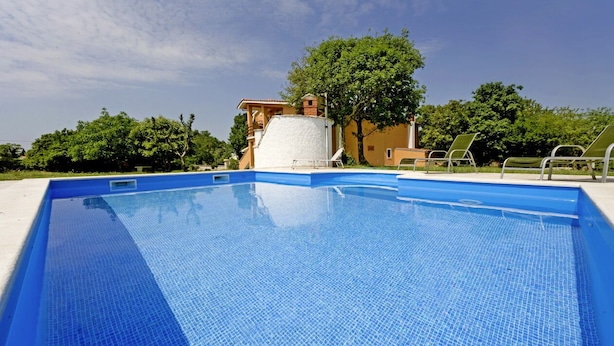 Ein Schwimmbecken mit Fliesenrand, ein kleines Gebäude mit rotem Dach und ein Baum im Hintergrund.
