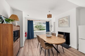 A modern kitchen with a wooden dining table, chairs, a hanging light fixture, and a window with curtains.