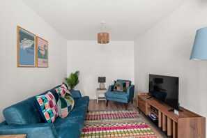 A modern living room with a blue sofa, a wooden TV stand, a TV, a patterned rug, and two framed artworks on the wall.