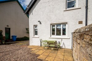 Ein weißes Gebäude mit einer Terrassenfläche, einem Tisch mit Stühlen und einer blauen Mülltonne.