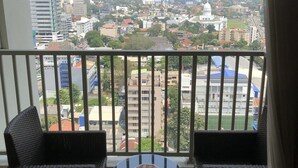 Un balcón con dos sillas de mimbre y una mesa pequeña, con vista a un paisaje urbano que incluye varios edificios y un campo deportivo.