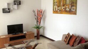 Una sala de estar moderna con televisor de pantalla plana, mueble de entretenimiento de madera, mesa de centro de vidrio y un sofá color beige con cojines estampados.