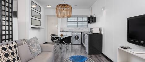 Una sala de estar moderna con un sofá, una mesa de centro y un televisor. Hay una zona de cocina con lavadora y una mesa de comedor.