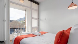 Una camera d'albergo con un letto, una sedia bianca e un balcone che si affaccia su edifici e montagne.