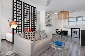 A modern living room with a sofa, a coffee table, and a floor lamp. There is a kitchen area with a washing machine and a dining table with chairs. - Cascades 508 (Cape Town)