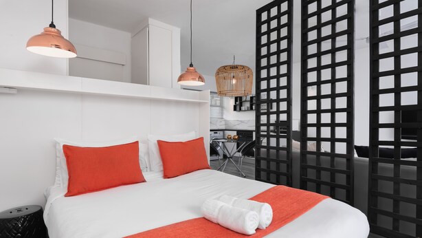 Una camera d'albergo moderna con un letto grande, cuscini arancioni, una coperta bianca e una coperta colorata rossa. Sono presenti due lampade a sospensione e un cesto di vimini appeso al soffitto.