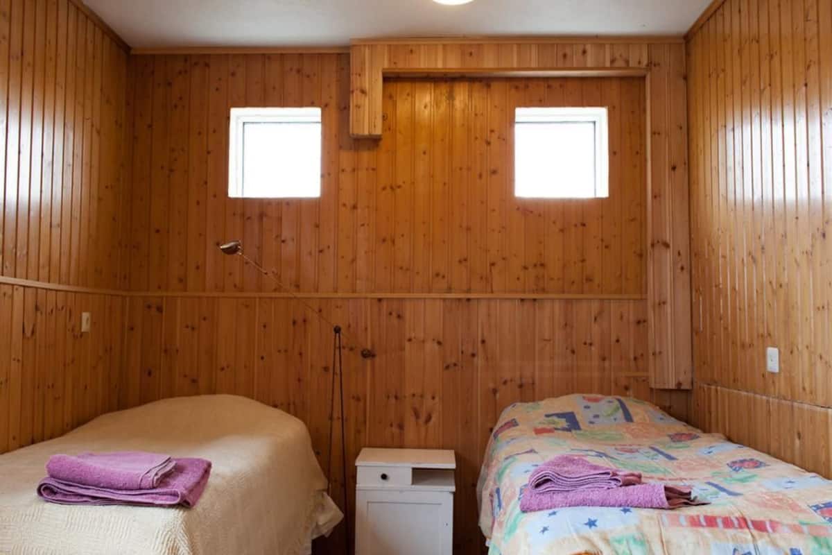 Una habitación con paredes de madera, dos camas, un armario pequeño blanco y dos ventanas.