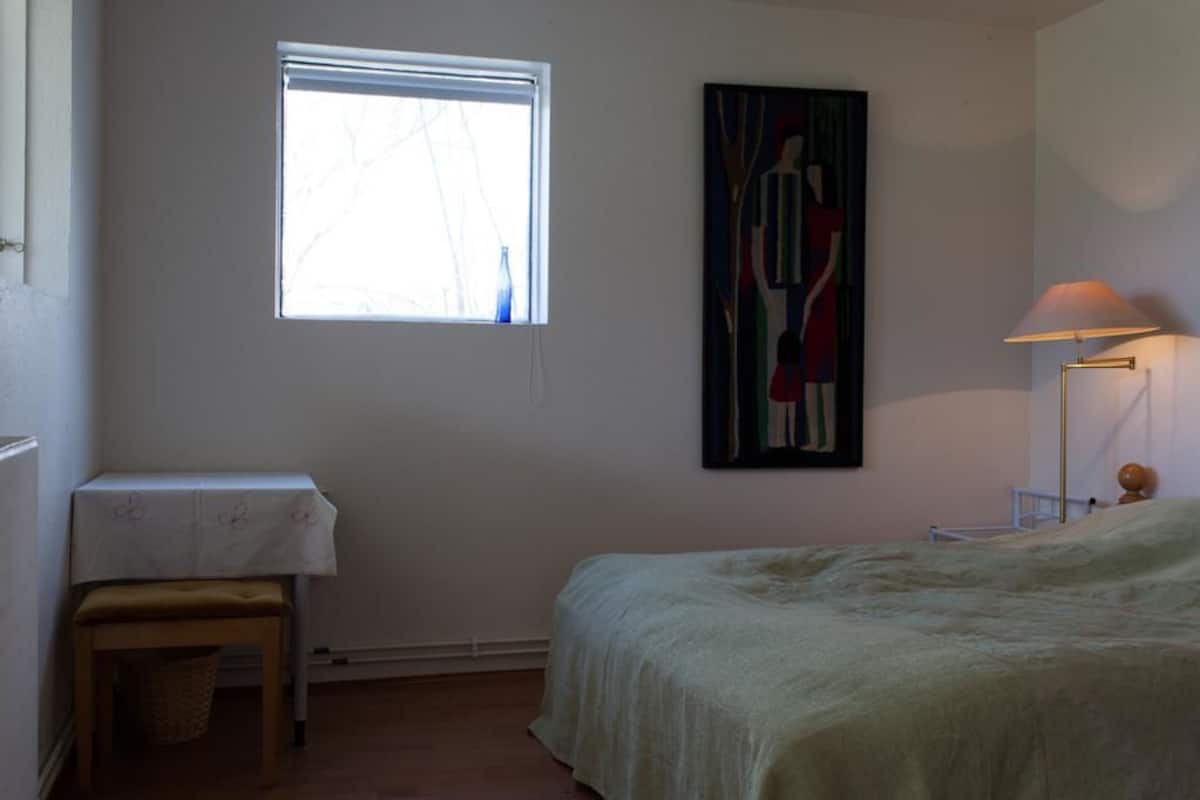 Un dormitorio con cama, ventana, mesa con mantel, lámpara y un cuadro en la pared.