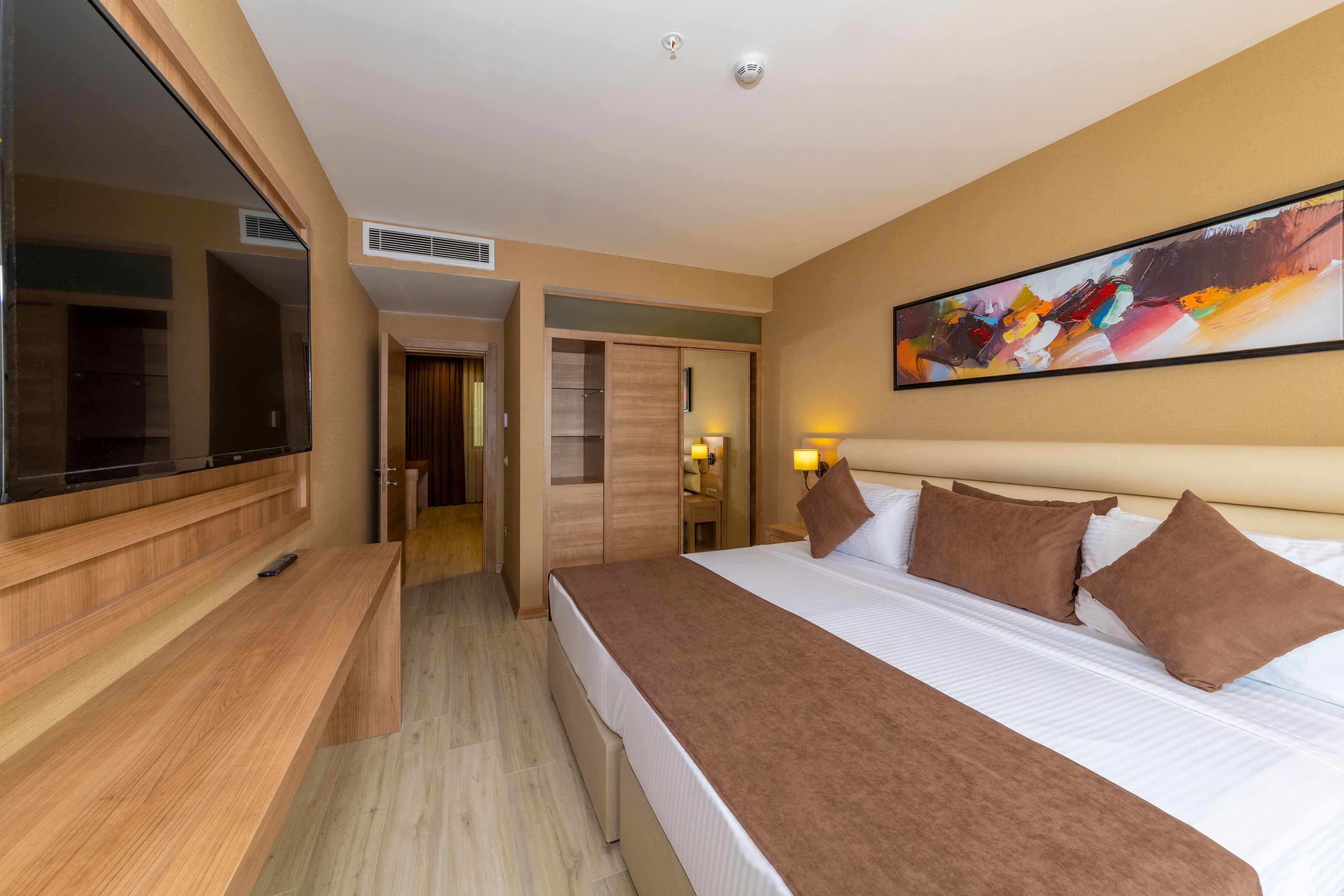 A modern hotel room with a large bed, a flat-screen TV, a wooden desk, and a painting on the wall.