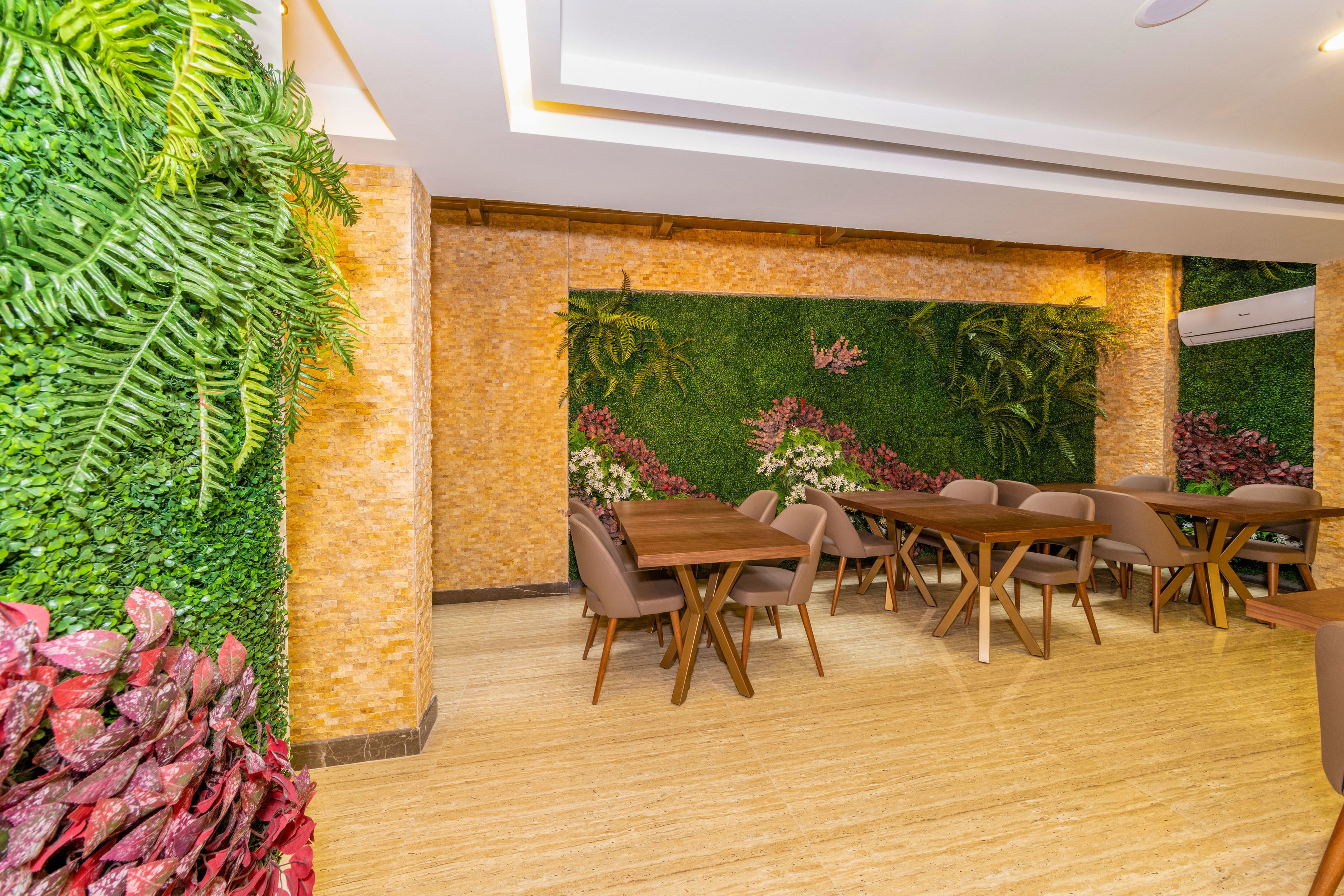 A dining area with wooden tables and chairs, a green wall with plants, and a tiled floor.