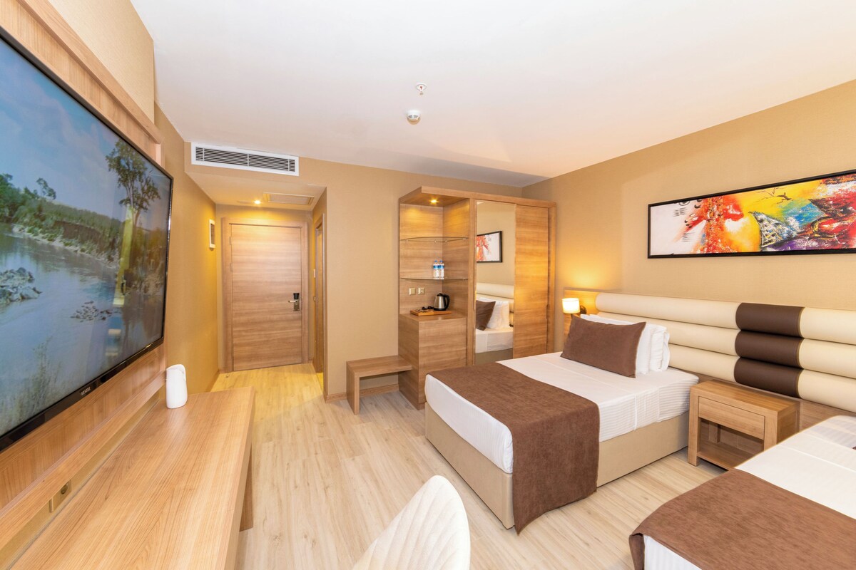 Une chambre d’hôtel avec une grande télévision, un lit recouvert d’une couverture marron, une table de chevet et un tableau accroché au mur.