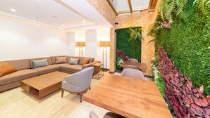 Un salon moderne avec un canapé d’angle, une table basse en bois et une table à manger. Un mur vert, agrémenté de plantes, se trouve derrière le coin salon, et une grande fenêtre surplombe cet espace.