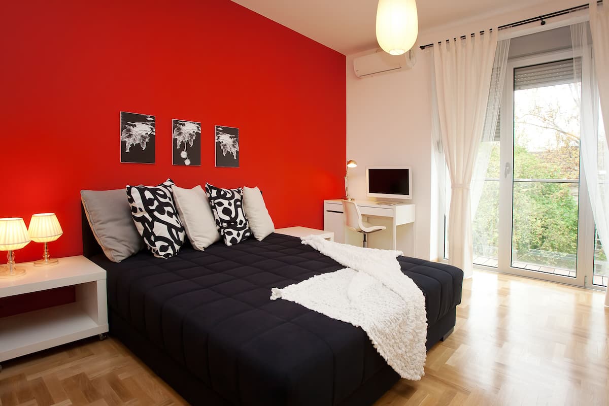 A modern bedroom with a red accent wall, a large bed with black and white bedding, a desk with a computer, and a large window with curtains.