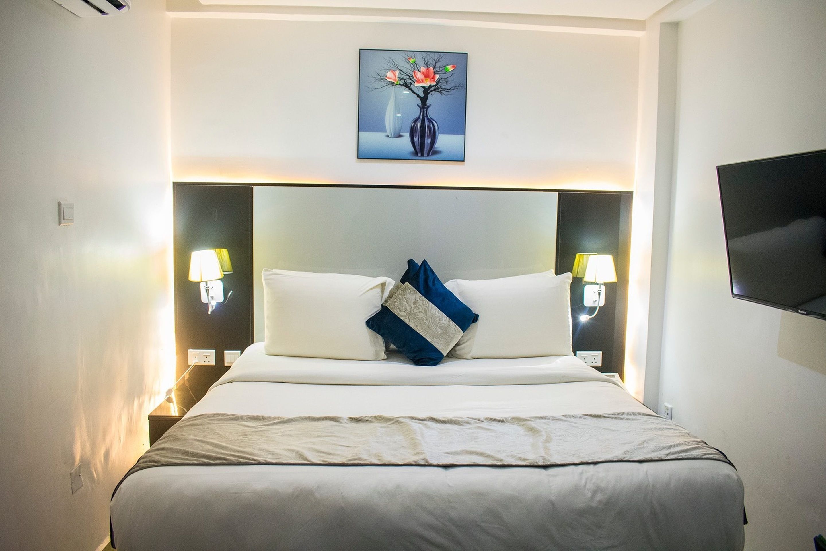 A hotel room with a large bed, two bedside lamps, a flat-screen TV, and a painting above the bed.