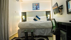 A hotel room with a bed, bedside lamps, a desk, a chair, a TV, and a painting on the wall. - Ziroc Residence (Lekki)