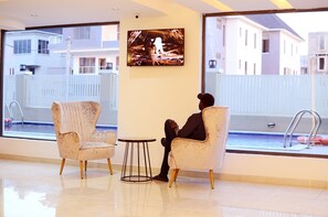 Quarto de hotel moderno com uma janela grande, uma televisão, duas poltronas e uma mesa redonda pequena.