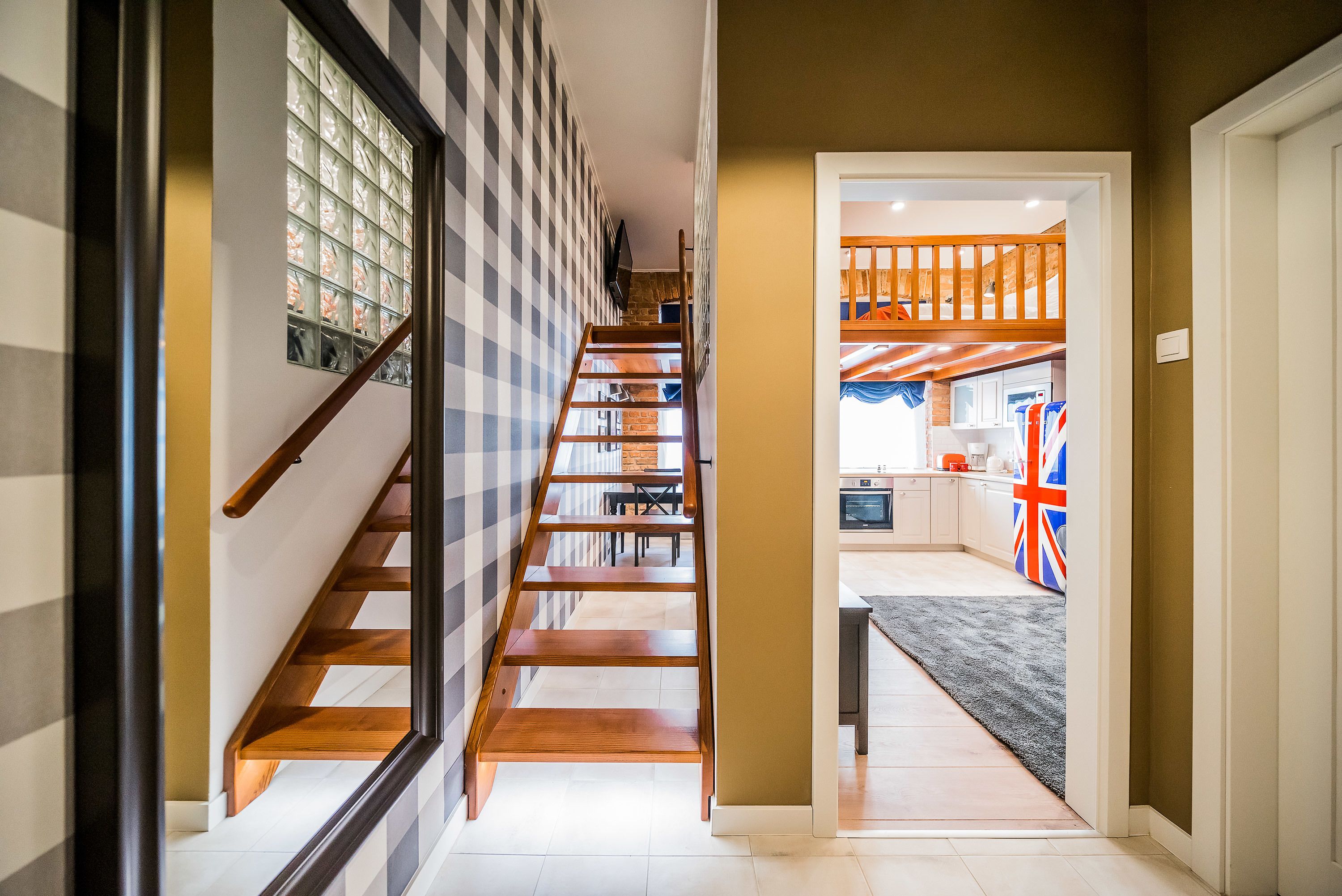 Un escalier menant à une cuisine, orné d’un drapeau Union Jack, d’un mur à carreaux et d’une main courante en bois.