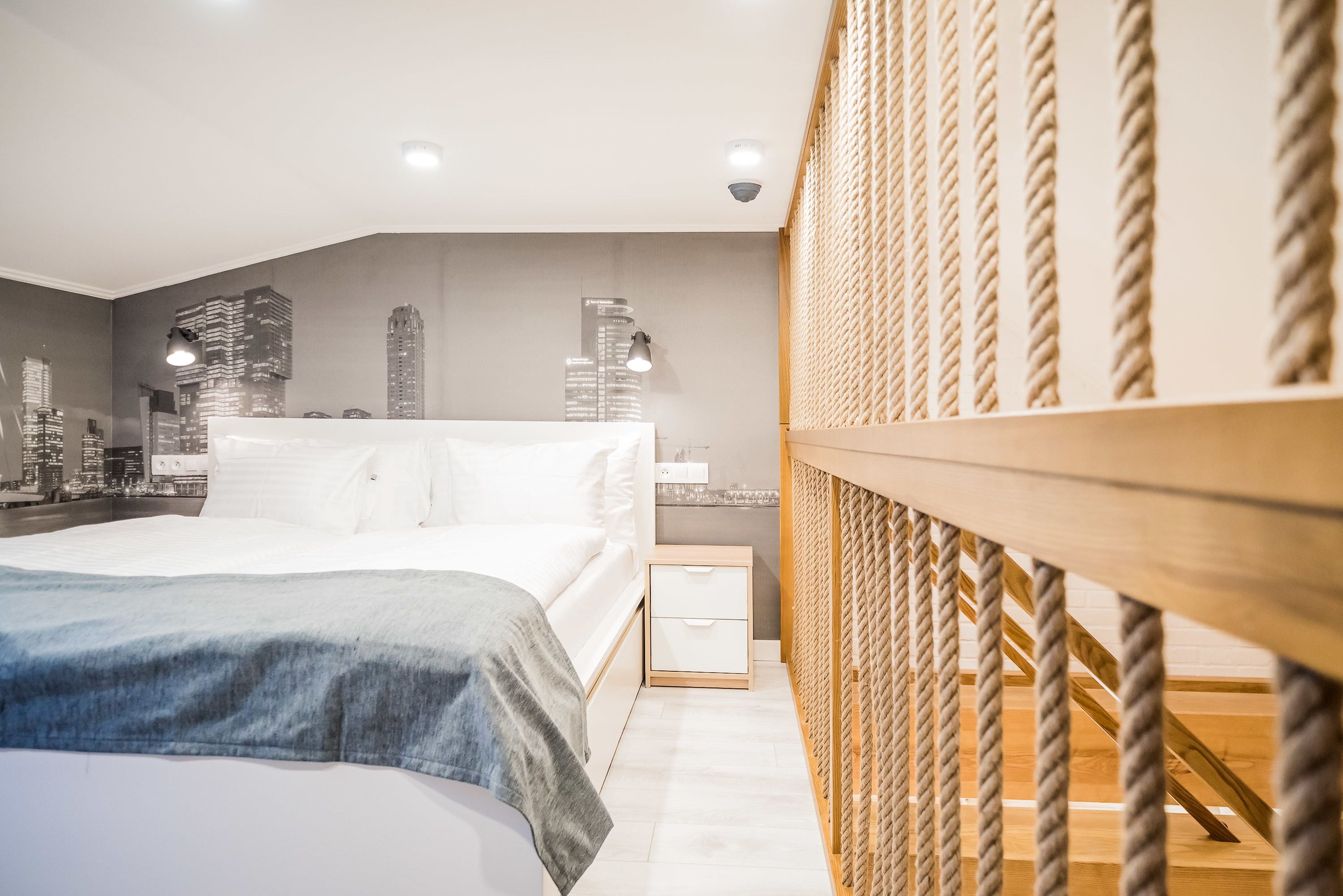 A modern bedroom with a cityscape mural, a bed with a blue blanket, a wooden headboard, and a nightstand with a drawer.