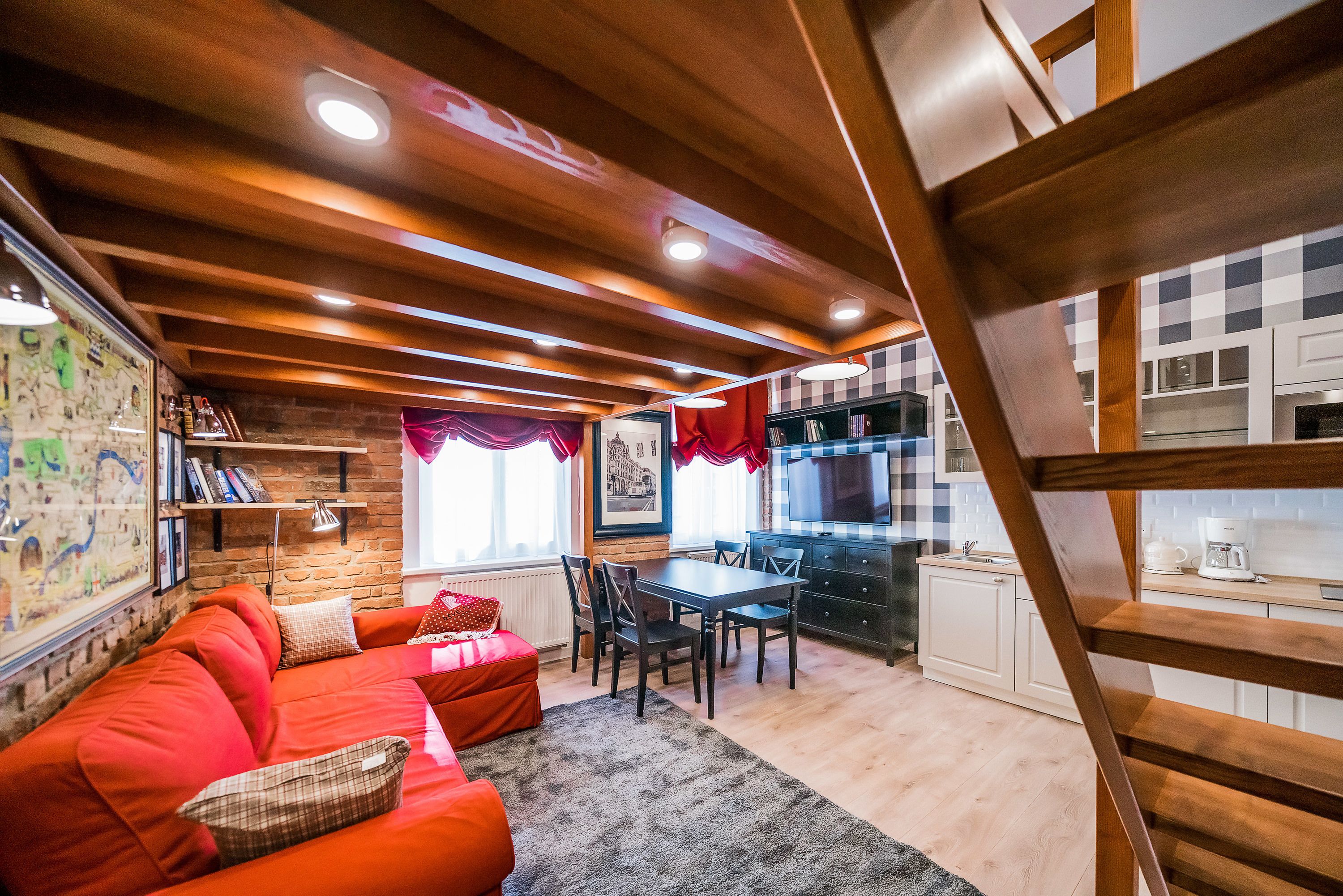 A cozy living room with a red sofa, a dining table, and a kitchen area with white cabinets and a wooden staircase.