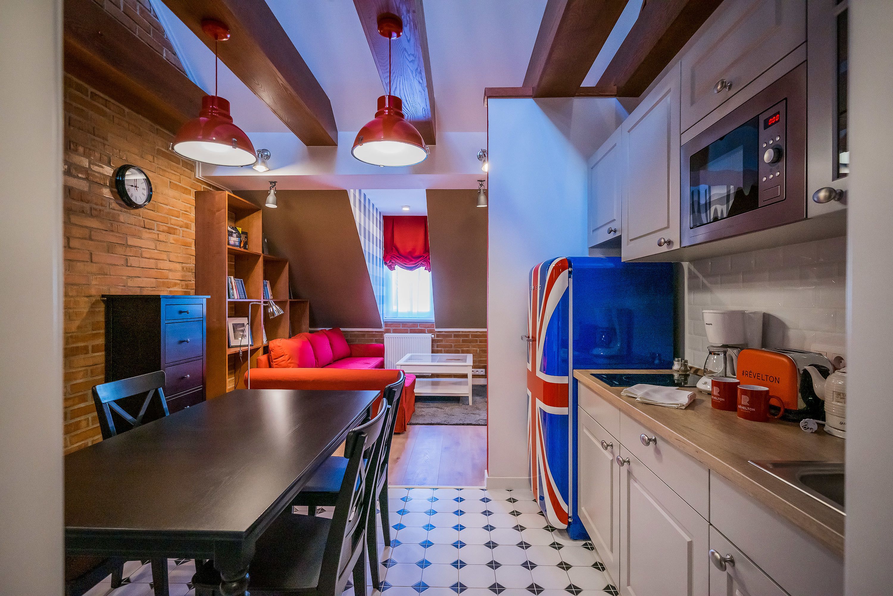 A compact kitchen with a microwave, toaster, and coffee maker. There is a dining area with a table and chairs, and a red sofa in the background.