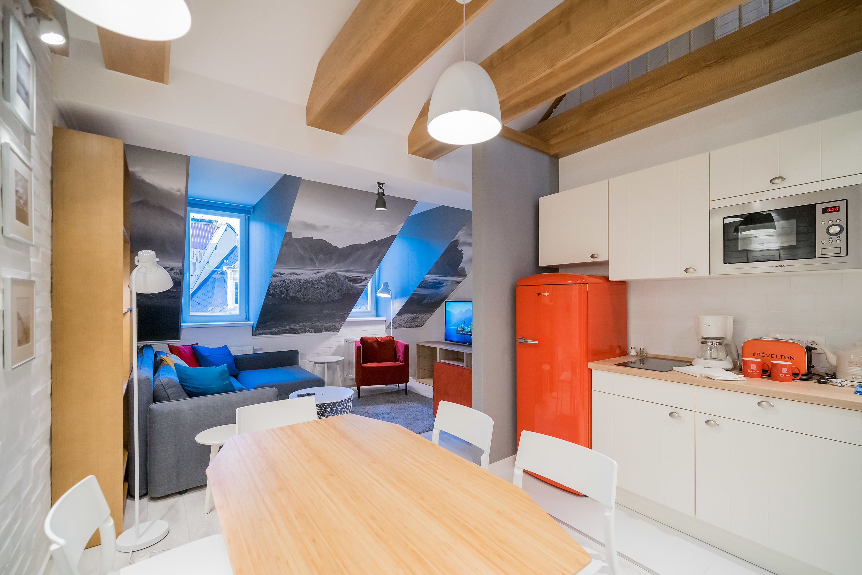 A modern kitchen with white cabinets, a microwave, and a coffee maker. There is a dining table with chairs and a sofa with blue cushions.