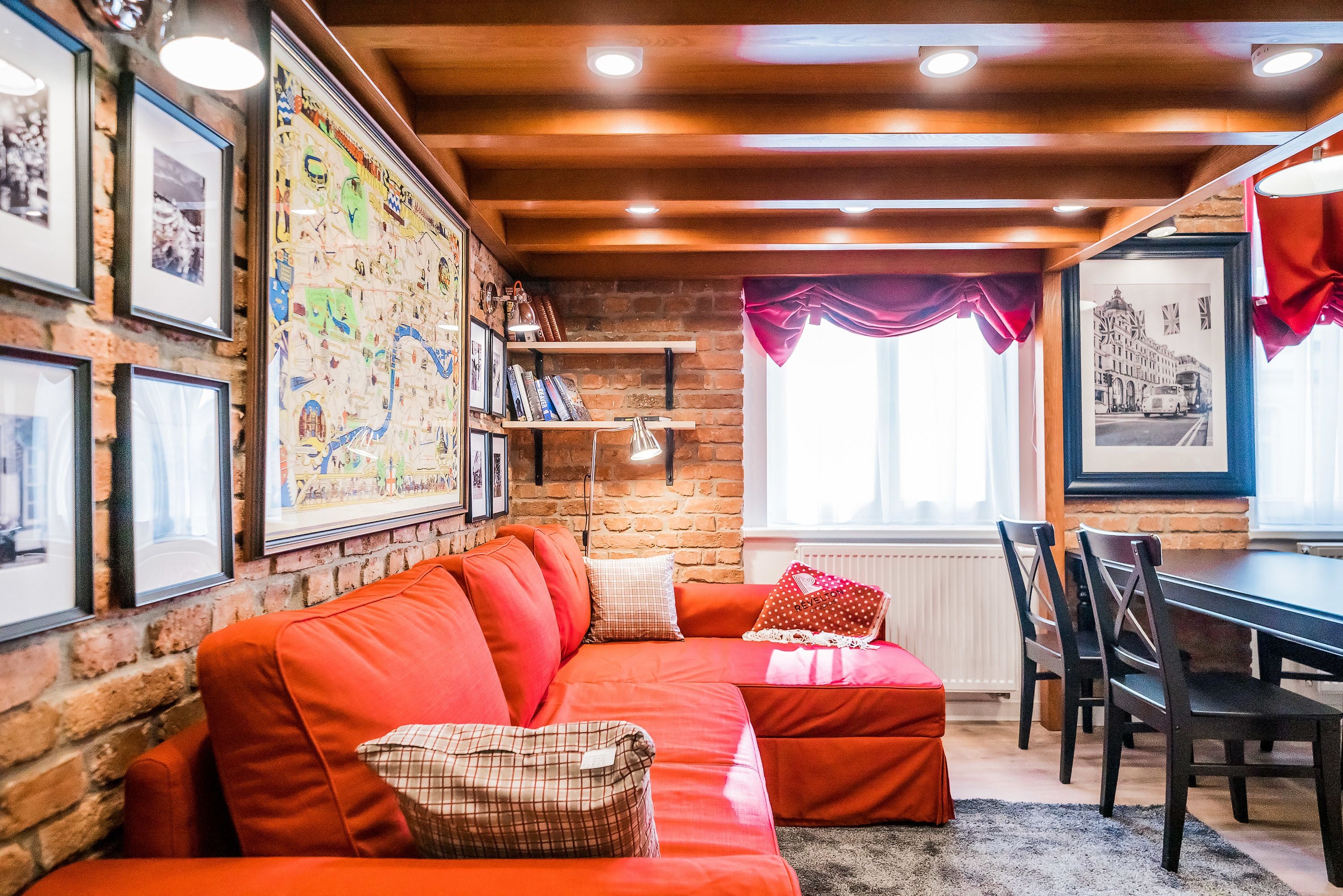 Ein gemütliches Zimmer mit Backsteinwand, Holzboden und roten Sofa, einem Esstisch mit Stühlen sowie gerahmten Bildern an den Wänden.