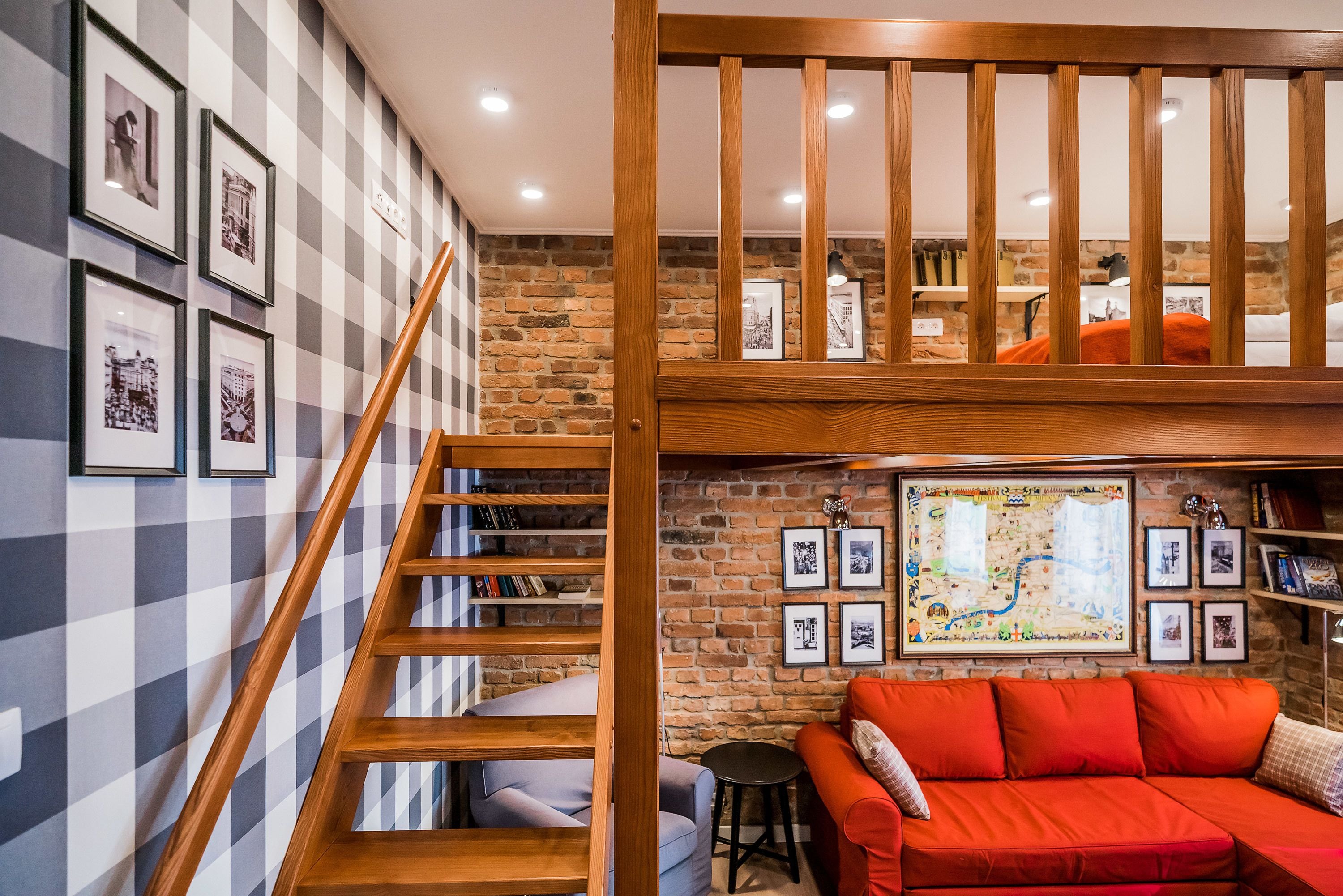 Una habitación estilo loft con escalera de madera, sofá rojo y pared de ladrillo con cuadros enmarcados.