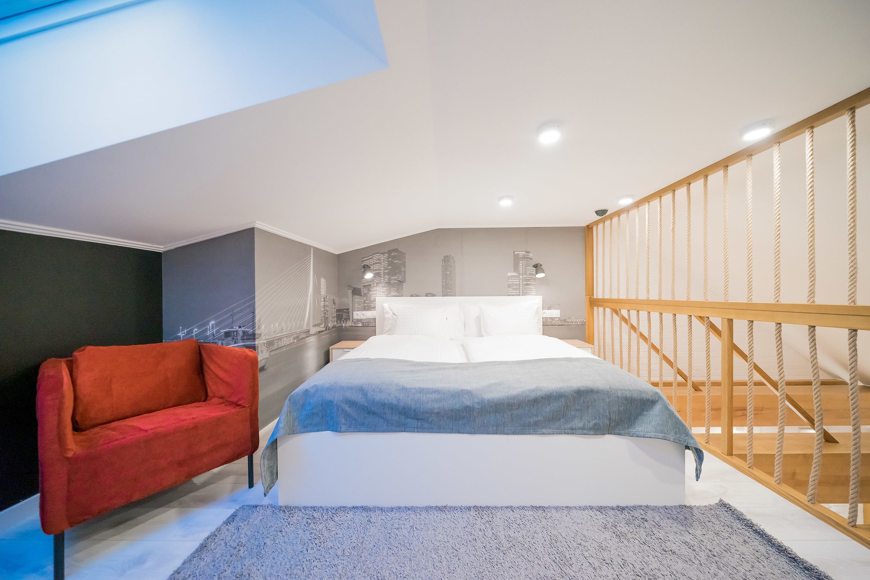 A modern bedroom with a bed, a red chair, a wooden staircase, and a cityscape mural on the wall.