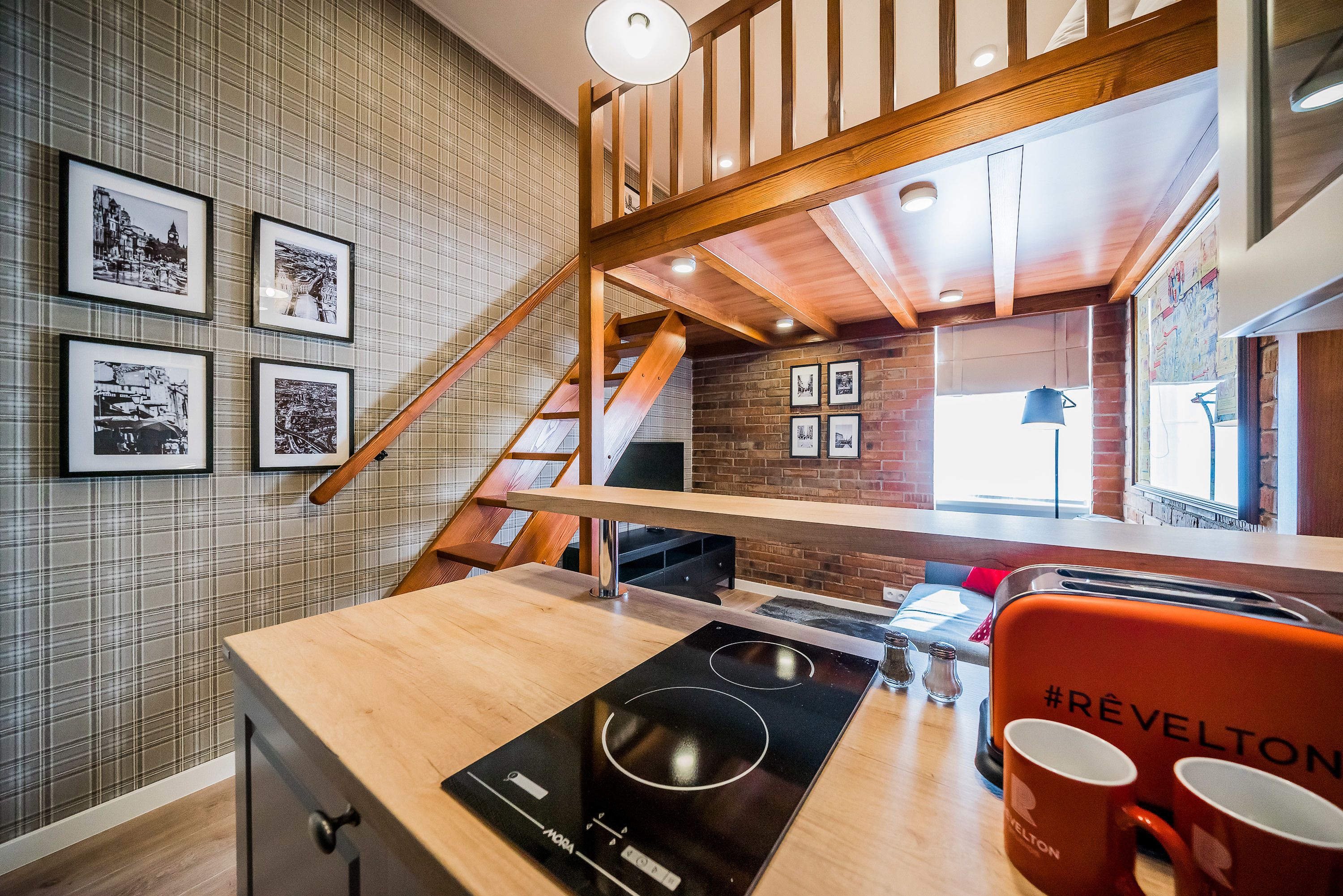 Cocina con encimera de madera, estufa integrada y una silla roja con el nombre de marca. En la pared hay cuadros enmarcados y una escalera que lleva a un nivel superior.
