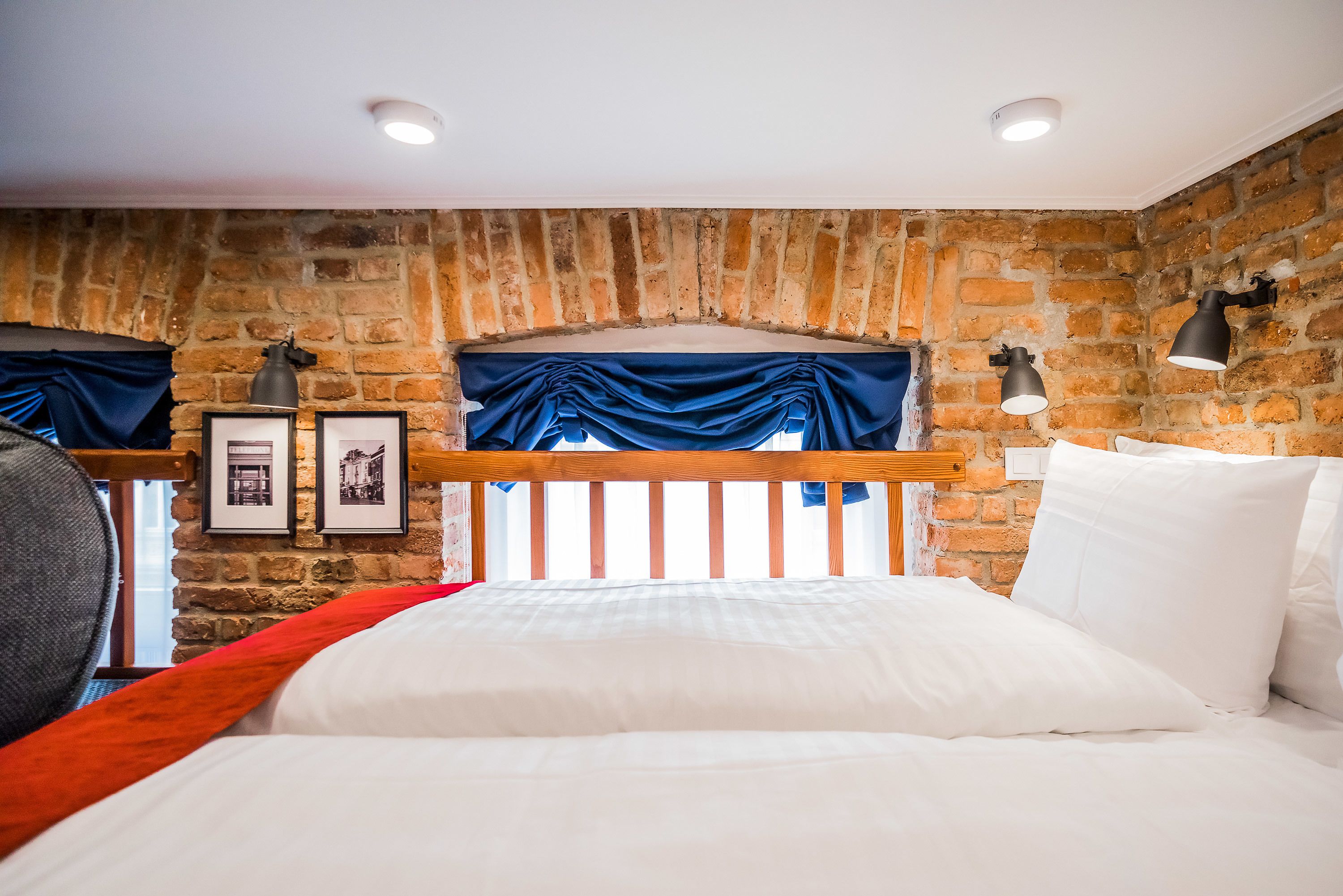 A bedroom with a brick wall, a wooden headboard, a bed with white bedding, two framed pictures, and two wall-mounted lamps.