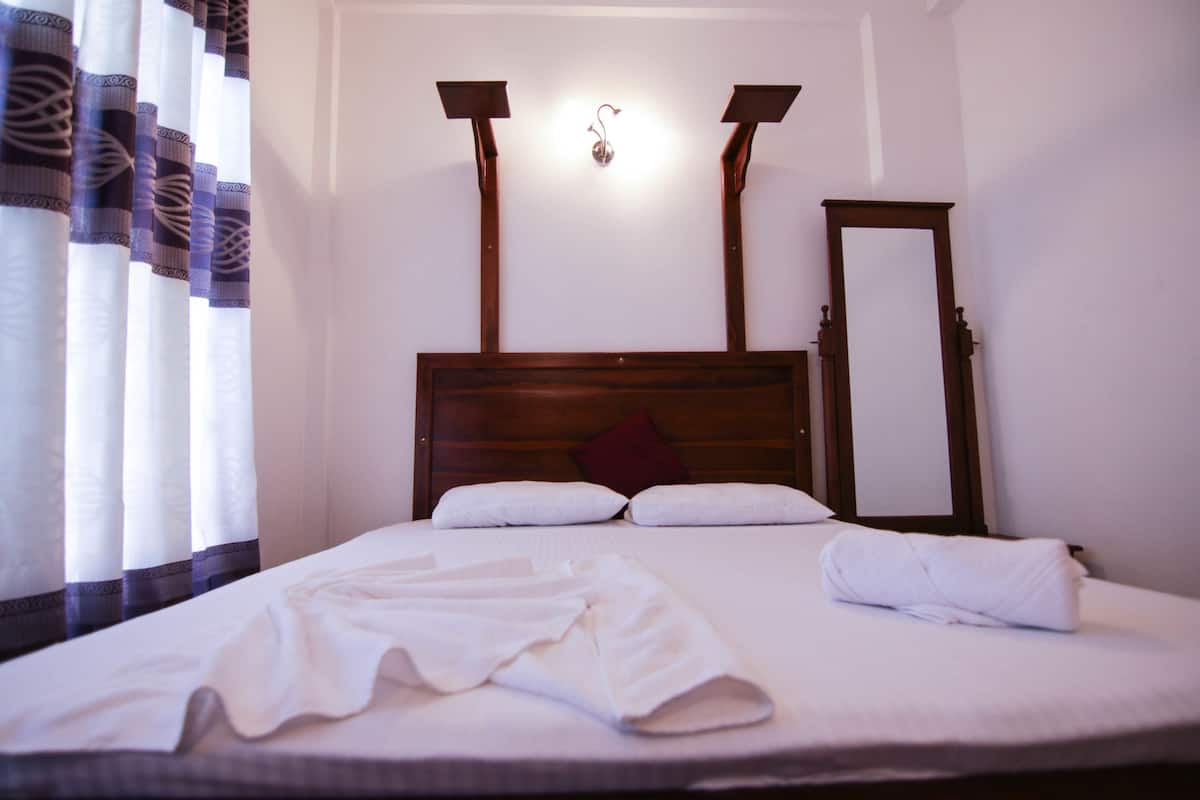 A neatly made bed with white linens and a red pillow, a wooden headboard, and a mirror on the wall.