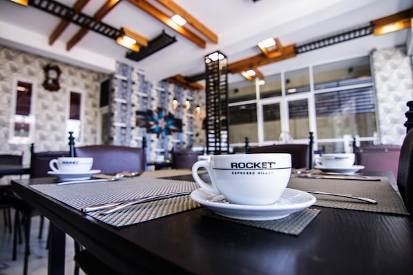 A coffee cup labeled "ROCKET ESPRESSO MILANO" on a table in a restaurant with patterned wallpaper and wooden beams.