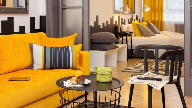 Ein modernes Wohnzimmer mit einem gelben Sofa, einem runden Couchtisch mit einer Zeitung, einem schwarzen Sessel und einem Bett mit gelben Vorhängen.
