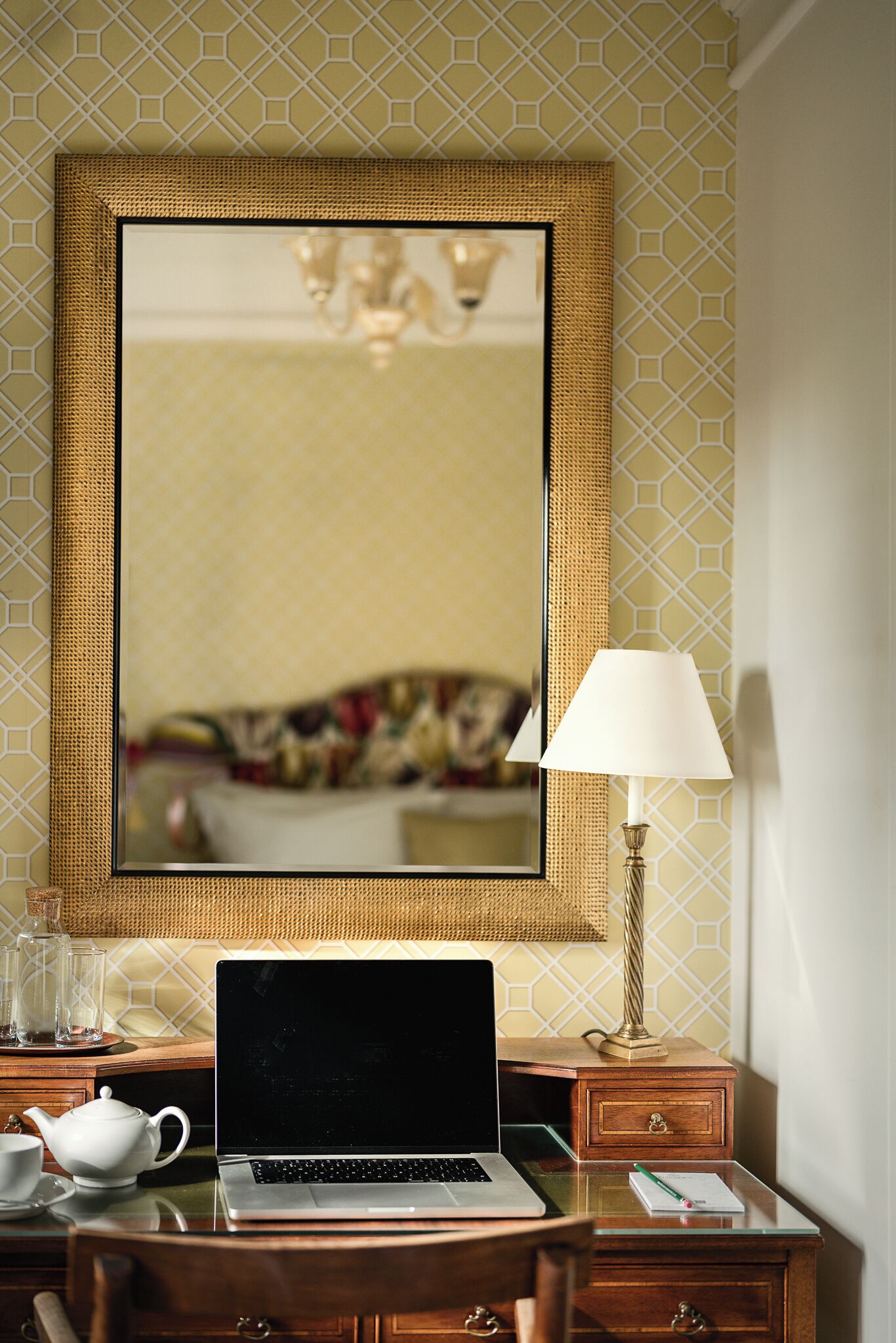 A hotel room with a laptop on a desk, a lamp, a teapot, and a mirror reflecting a bed and chandelier.