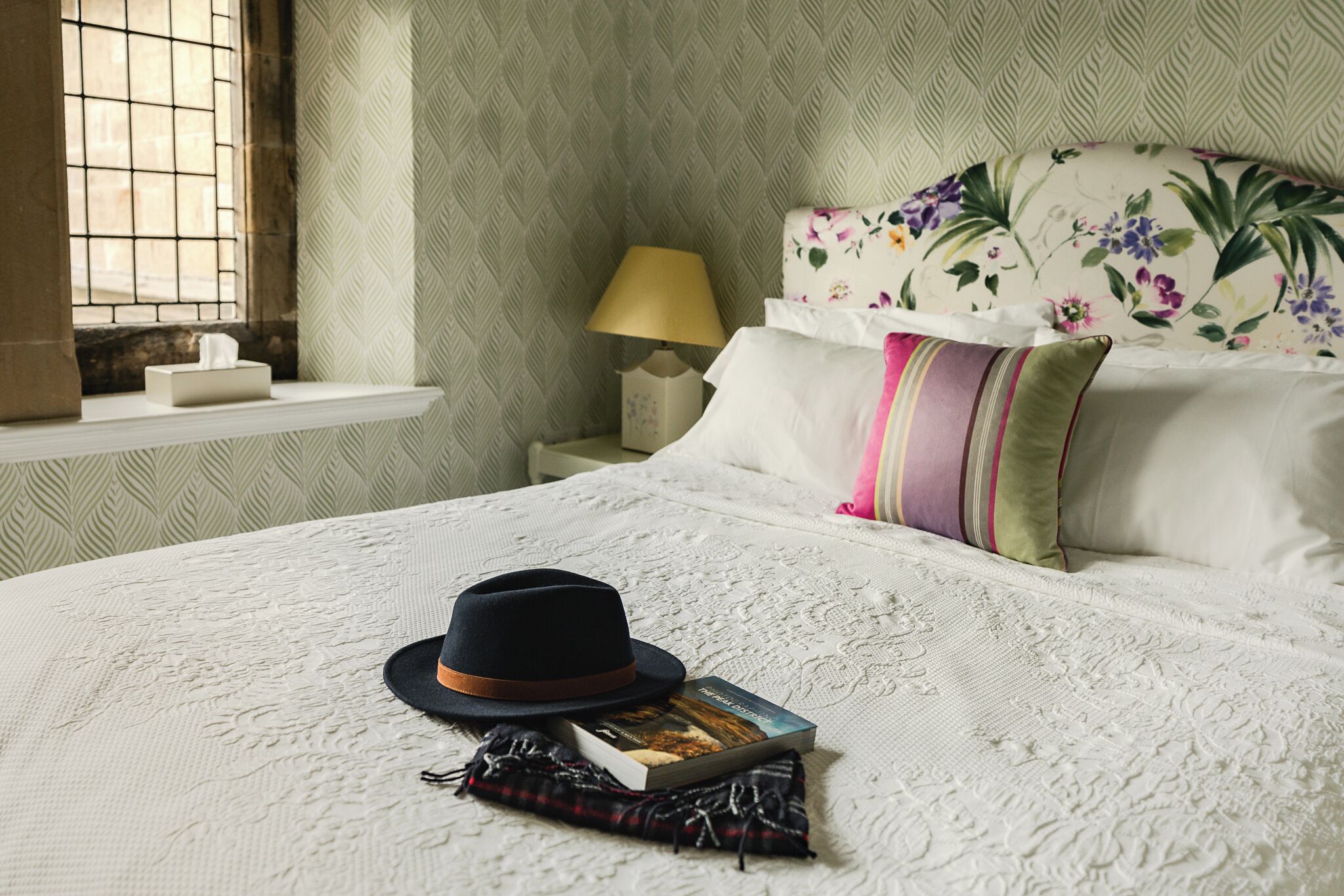 Een net opgemaakt bed met een boek en een hoed, een bloemen hoofdbord, een bedlampje en een raam met tralies.