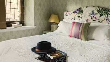 Een net opgemaakt bed met een boek en een hoed, een bloemen hoofdbord, een bedlampje en een raam met tralies.
