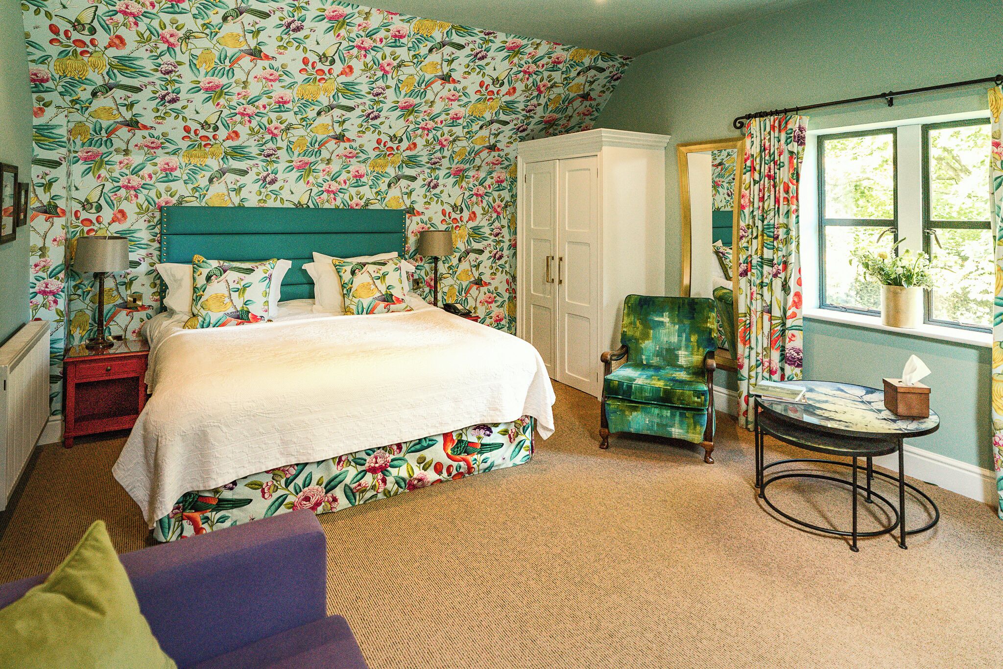 A bedroom with a floral wallpaper, a bed with a white bedspread, a green armchair, a small table with a tissue box, and a white wardrobe.
