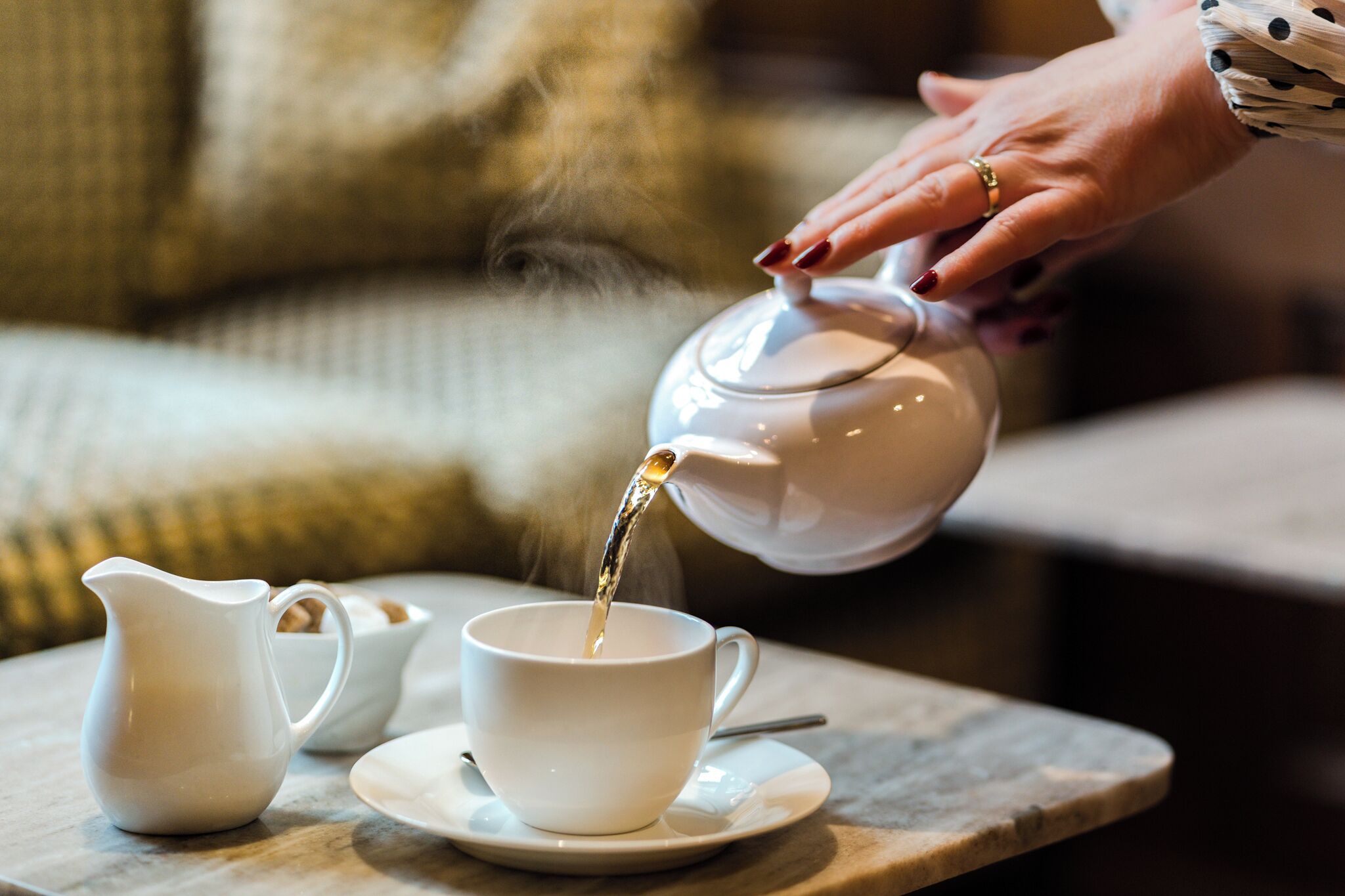 A white teapot pouring tea into a white cup on a table with a saucer, a smaller white pitcher, and a spoon.