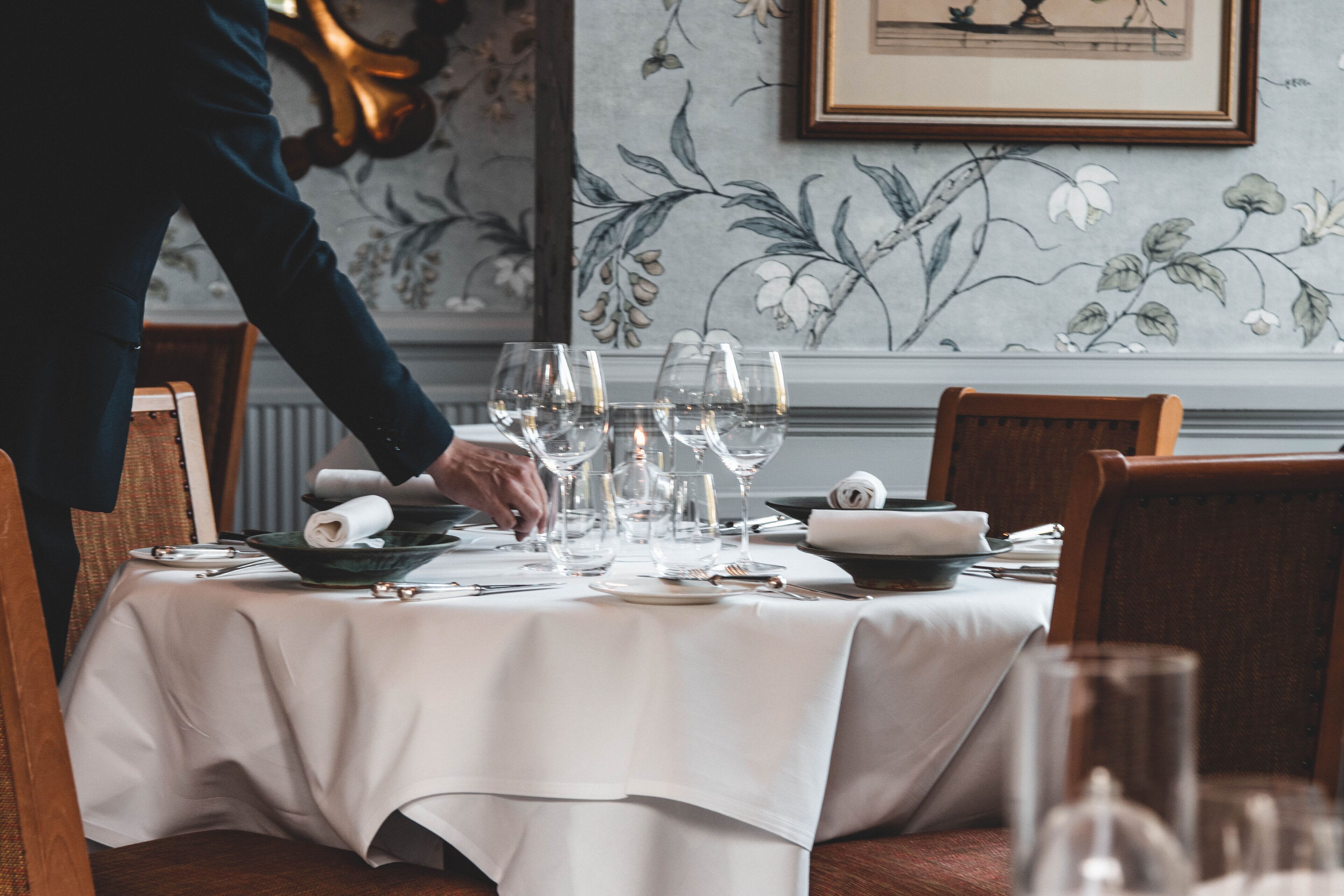 A dining table set with wine glasses, plates, and cutlery, a person in a suit, and floral wallpaper.