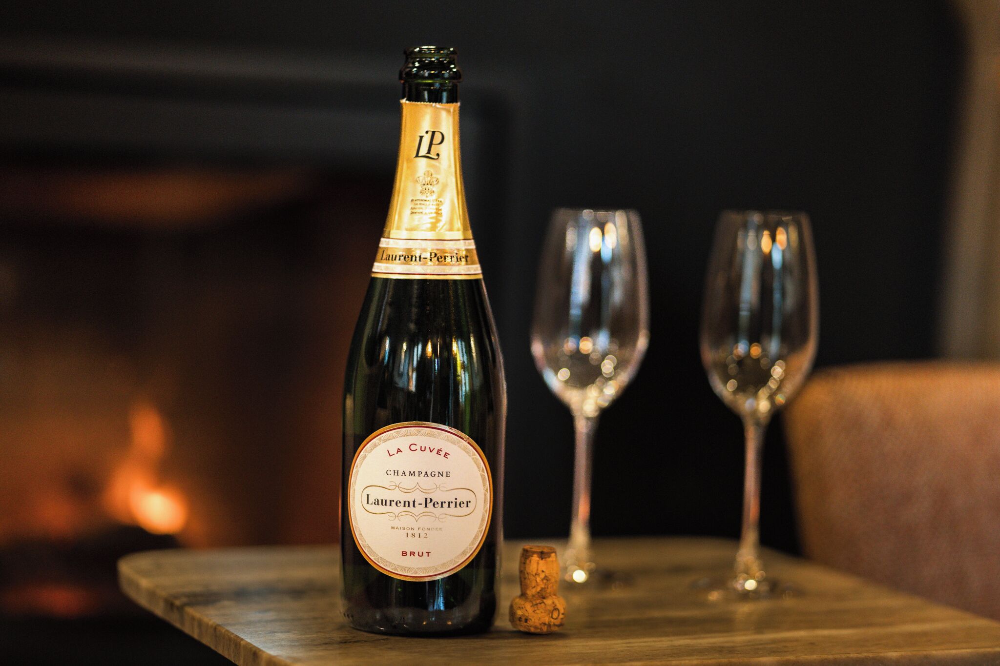 A bottle of champagne, a cork, and two glasses on a wooden table.