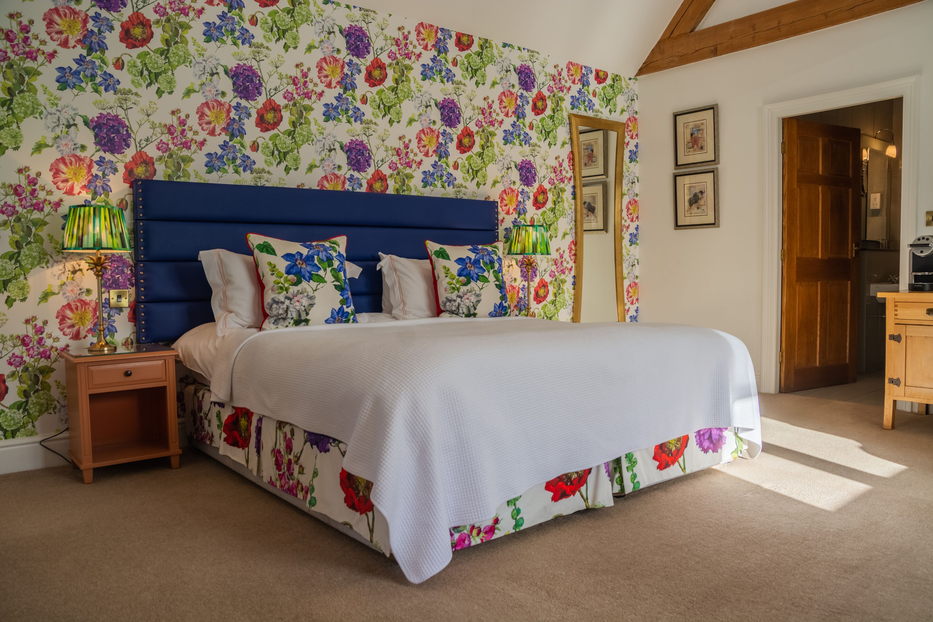 A bedroom with a floral wallpaper, a blue headboard, a white bedspread, and a wooden nightstand with a lamp.